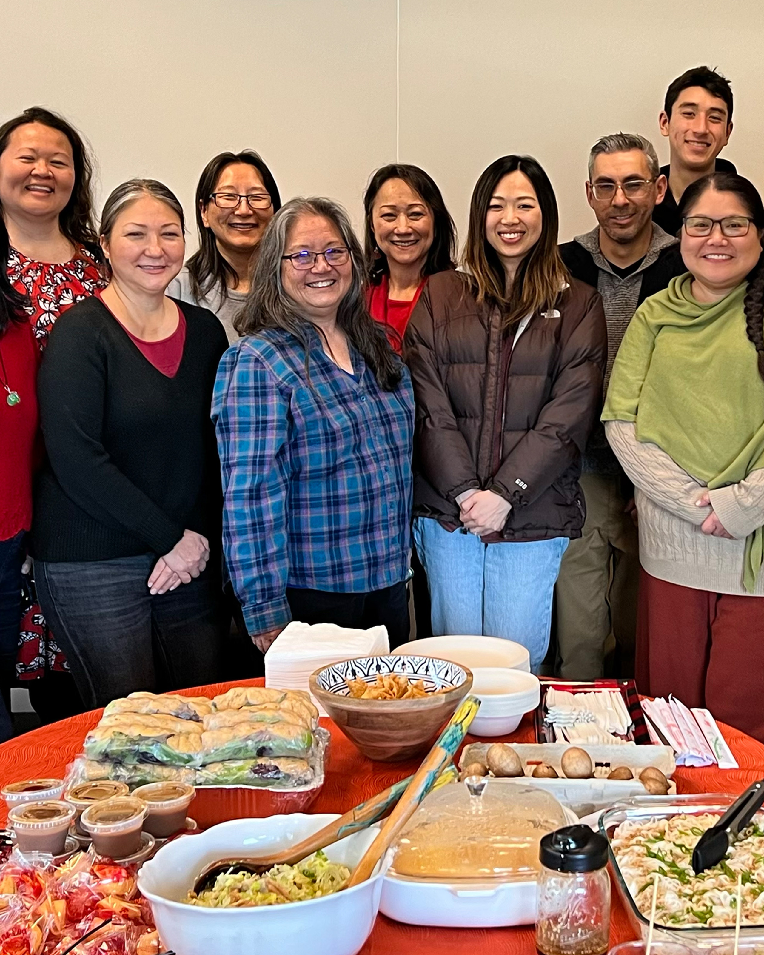Group of people standing behind a table with dishes at a casual indoor gathering.